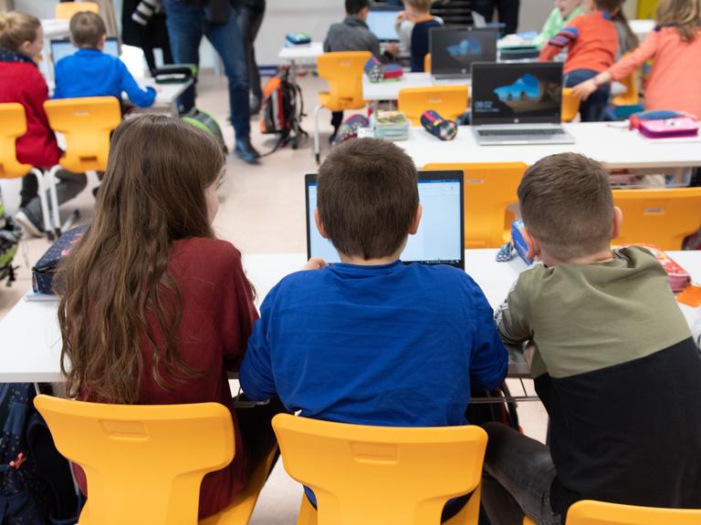 Drei Grundschüler sitzen in einem vollen Klassenzimmer vor einem Laptop.