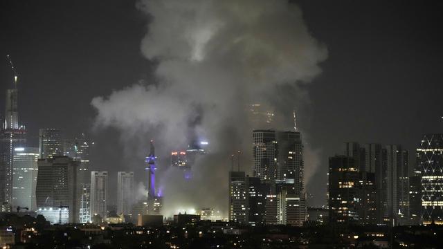 Es ist Nacht. Man sieht Gebäude und die Lichter von der Stadt Tel Aviv in Israel aus der Ferne. Dichter grauer Rauch steigt über Häusern auf.