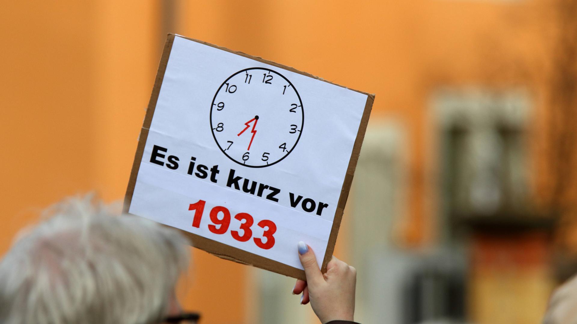 Ein Demonstrant hält ein Schild in die Höhe, auf dem ein Zifferblatt einer Uhr zu sehen ist und "Es ist kurz vor 1933" steht.