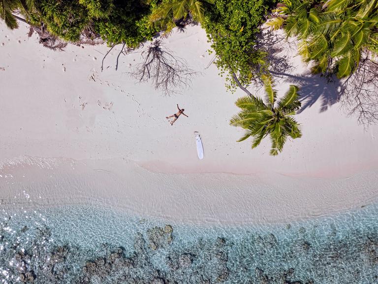 Ein Mann liegt mit einem Surfboard an einem einsamen Strand.