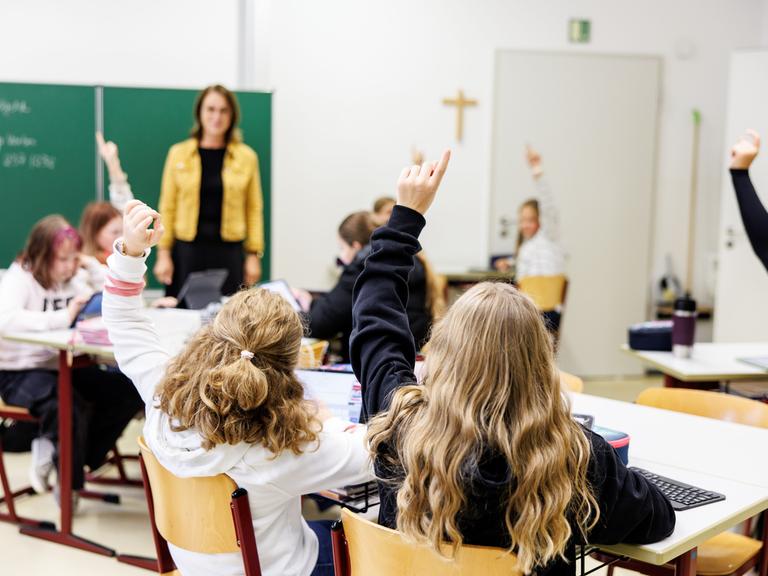 Zwei Schülerinnen zeigen in einer Klasse auf, dahinter steht eine Lehrerin vor der Tafel.