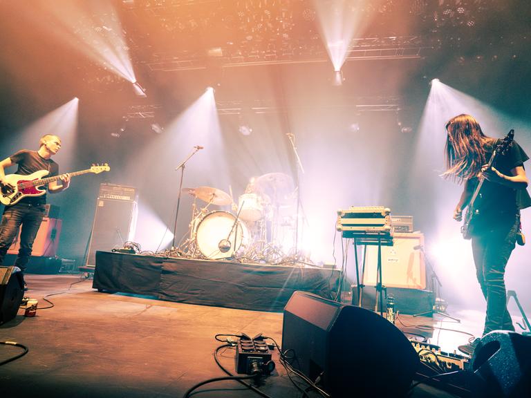 Eine Live-Rockband auf der Bühne, links ein Mann mit kurzen Haaren am E-Bass, rechts ein Mann mit langem Haar an der Gitarre, im Hintrergrund ein Schlagzeug. 