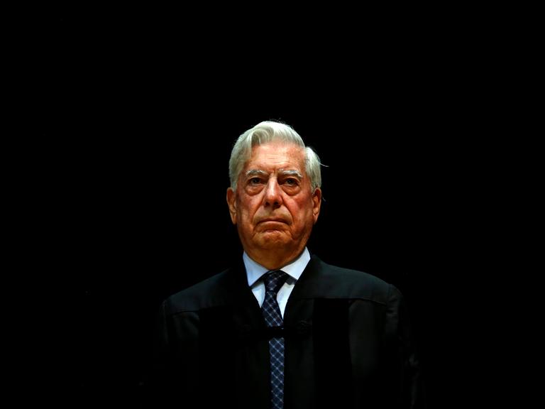 Mario Vargas Llosa steht vor einem schwarzen Hintergrund. Er trägt ein dunkles Sakko und eine blau-gemusterte Krawatte.