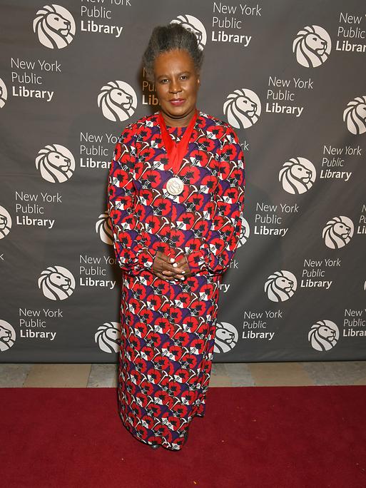 Claudia Rankine lächelt in die Kamera. Sie trägt ein langes Kleid mit rot-blauem Blumenmuster.