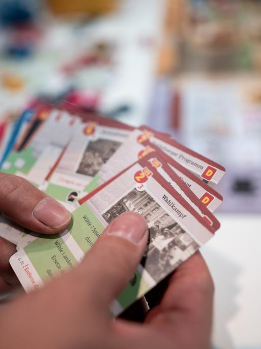Hände halten Karten in der Hand, im Hintergrund ist ein Tisch mit einem Spielbrett und Figuren.