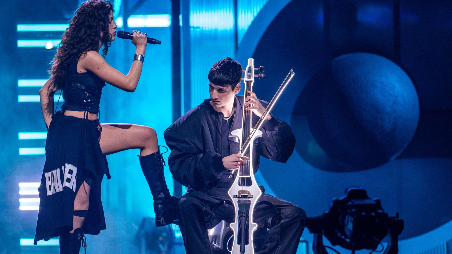 Das Duo Abor & Tynna während eines Auftritts. „Abor“ spielt Cello im Sitzen, während seine Schwester „Tynna“ stehend singt und ein Bein auf seinen Stuhl stellt. 