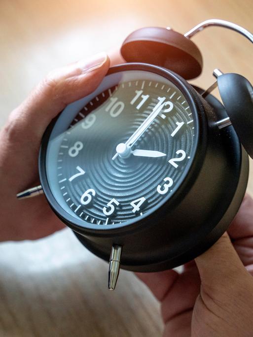 Zwei Hände drehen an einem Wecker, der auf 2 Uhr steht