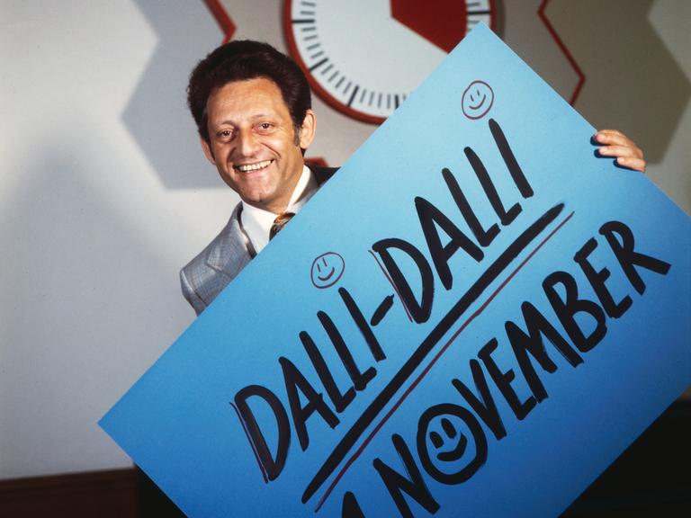 Hans Rosenthal vor der Stoppuhr in der Quiz- und Spielshow "Dalli-Dalli". Er hält ein Schild, auf dem steht: "Dalli Dalli 21. November"