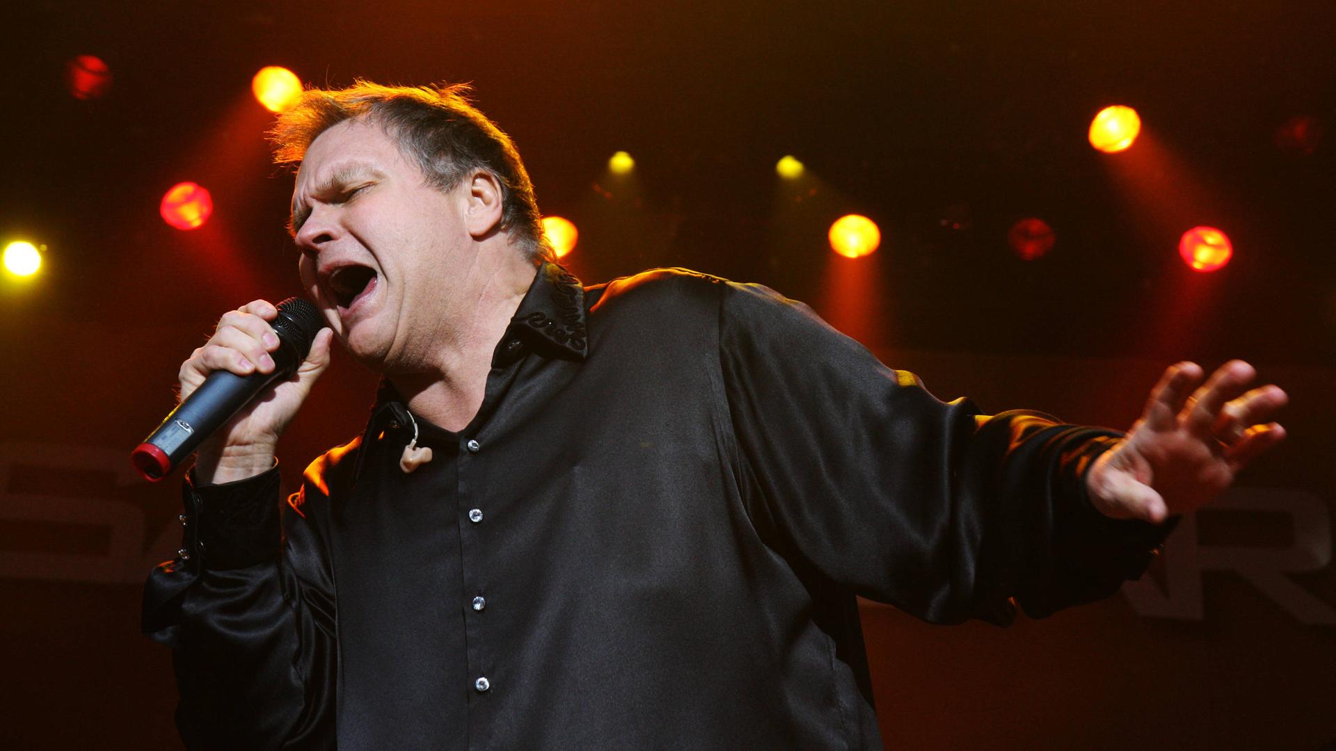 Ein Mann steht mit ausgebreiteten Armen auf einer Bühne und singt. In der rechten Hand hält er ein Mikrofon. Es ist der US-Rockmusiker Meat Loaf.