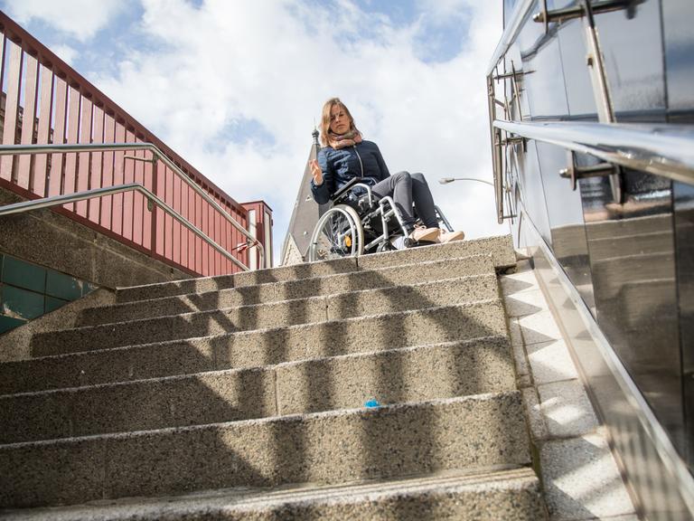 Eine Frau im Rollstuhl steht vor einer Treppe (gestellte Szene).
