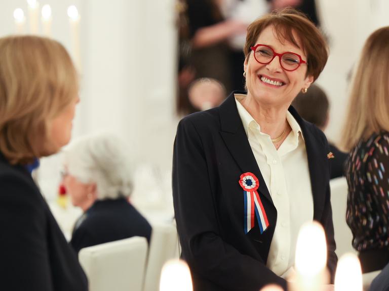 Hélène Miard-Delacroix trägt eine rote Brille und eine Brosche in den französischen Nationalfarben. Sie schaut zur Seite und lacht die Person links im Bild (Bundestagspräsidentin Bärbel Bas) an. 