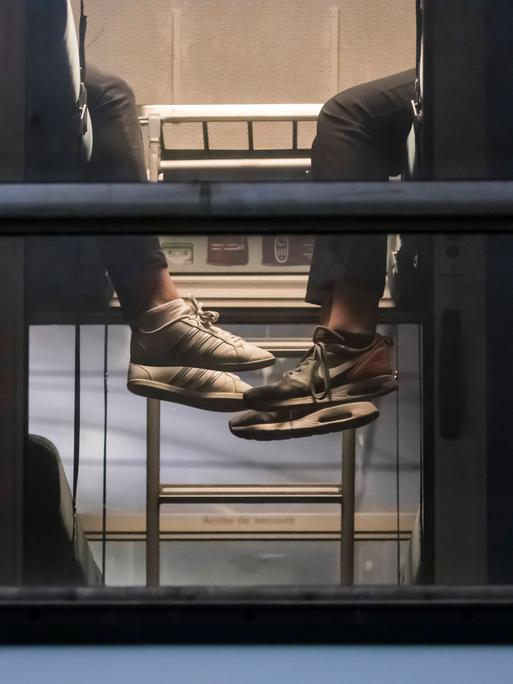 Blick von außen auf einen Schlafwagen. Zwei Personen, von denen nur Waden und Füße zu erkennen sind, sitzen sich gegenüber und lassen ihre Beine baumeln.