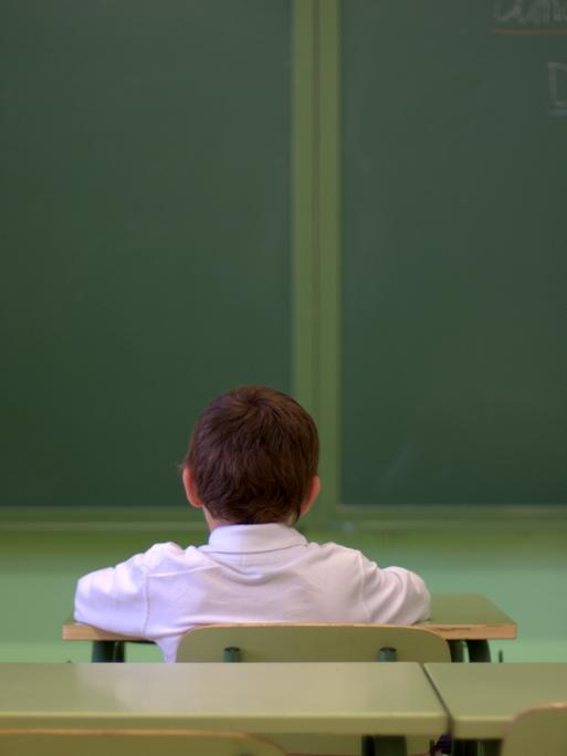 Ein Junge sitzt alleine in einem Klassenraum und guckt in Richtung der Tafel. Die Kamera hat ihn von hinten aufgenommen.
