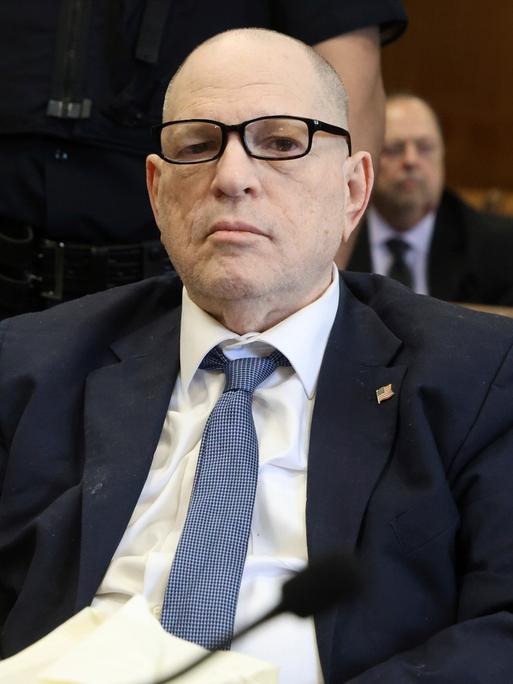 Ein älterer Mann mit Brille sitzt in einem Gerichtsaal und blickt ernst in die Kamera.