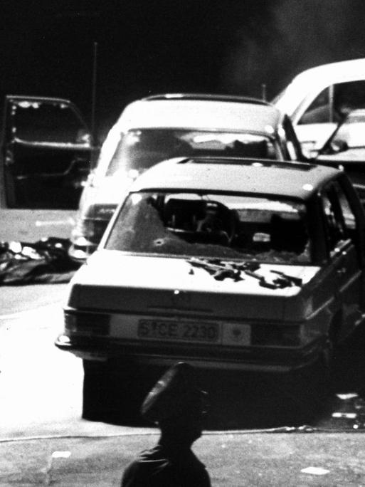Das Schwarz-Weiß-Foto zeigt drei stark beschädigte Autos, die im Dunkeln an einer Straße stehen. Teilweise sind die Scheiben zerstört, bei einem Auto steht die Motorhaube auf. Auf dem Boden sind abgedeckte Körper zu erahnen.
