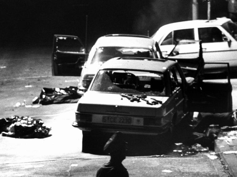 Das Schwarz-Weiß-Foto zeigt drei stark beschädigte Autos, die im Dunkeln an einer Straße stehen. Teilweise sind die Scheiben zerstört, bei einem Auto steht die Motorhaube auf. Auf dem Boden sind abgedeckte Körper zu erahnen.
