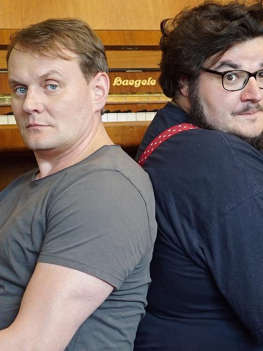 Devid Striesow und Axel Ranisch sitzen Rücken an Rücken vor einem Klavier.