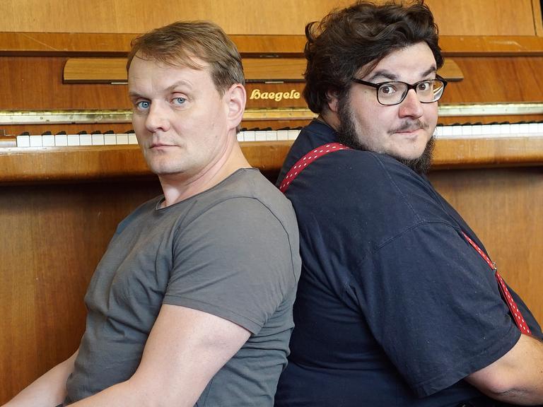 Devid Striesow und Axel Ranisch sitzen Rücken an Rücken vor einem Klavier.