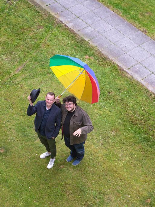 Devid Striesow und Axel Ranisch auf einer Wiese mit regenbogenfarbenen Regenschirm. Das Bild ist aus der Vogelperspektive fotografiert, beide schauen nach oben.