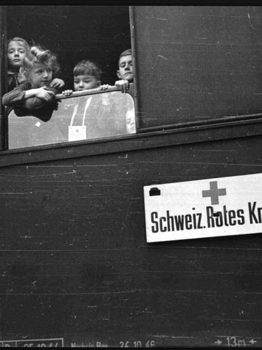 Schwarz-weiß-Foto: Kinder schauen aus einem Zugfenster. Am Zug ist ein Schild befestigt: "Schweizerisches Rotes Kreuz".