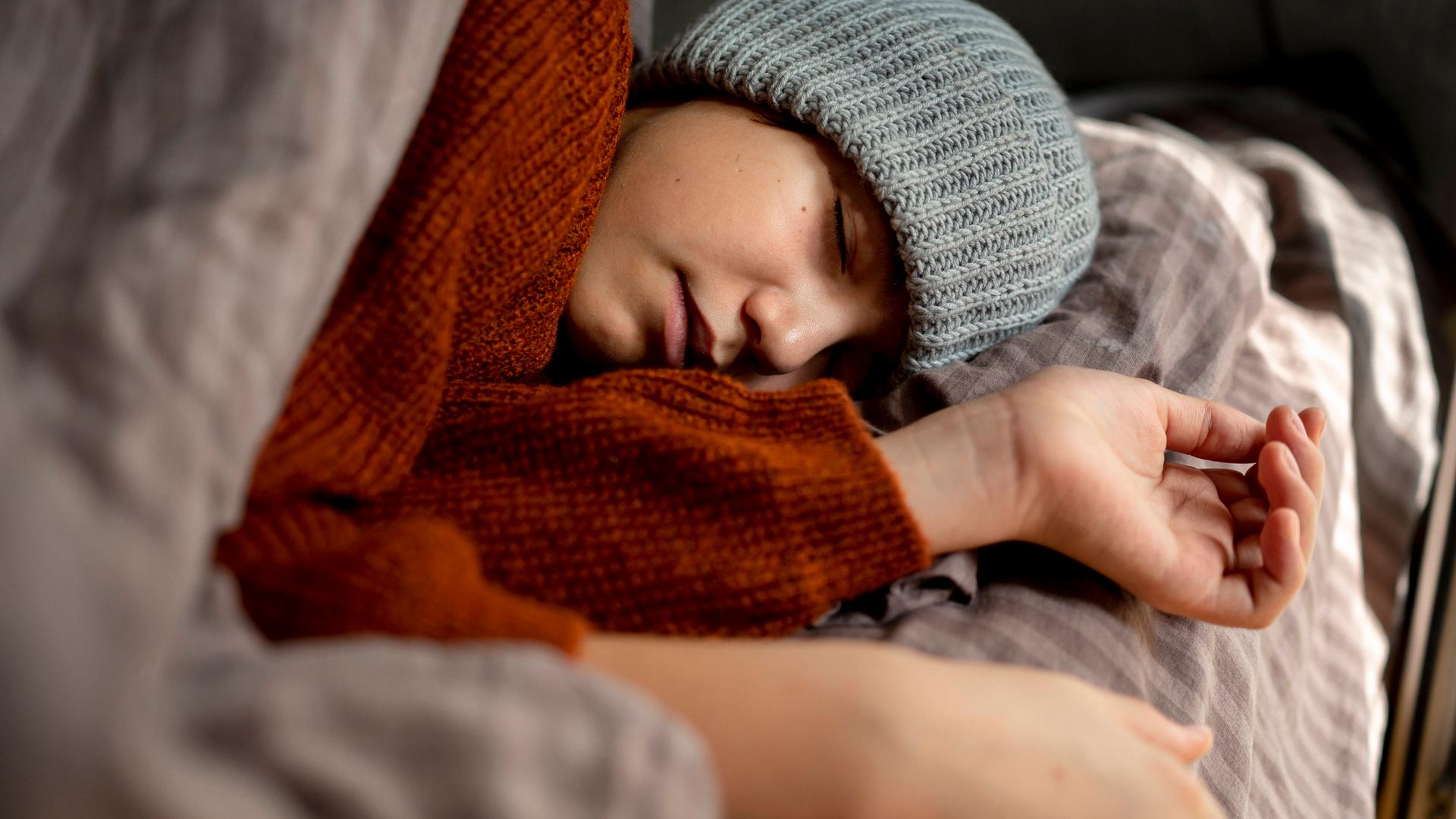 Ein Mensch mit Mütze schläft erschöpft in seinem Bett.(Symbolbild)