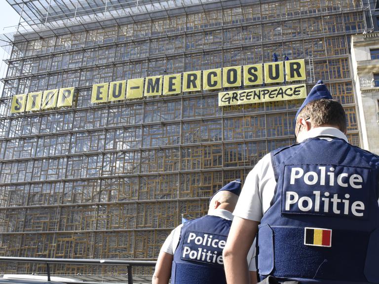 An einem Gebäude hängt ein langes gelbes Schild. Darauf steht in schwarzen Großbuchstaben: „Stop EU-Mercusor“. Darunter steht in kleinerer Schrift das Wort „Greenpeace“. 