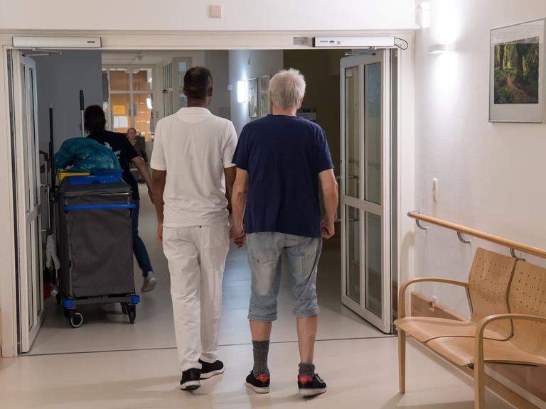 Ein pflegebedürftiger Mann (r) läuf mit einem Pfleger über den Flur.  