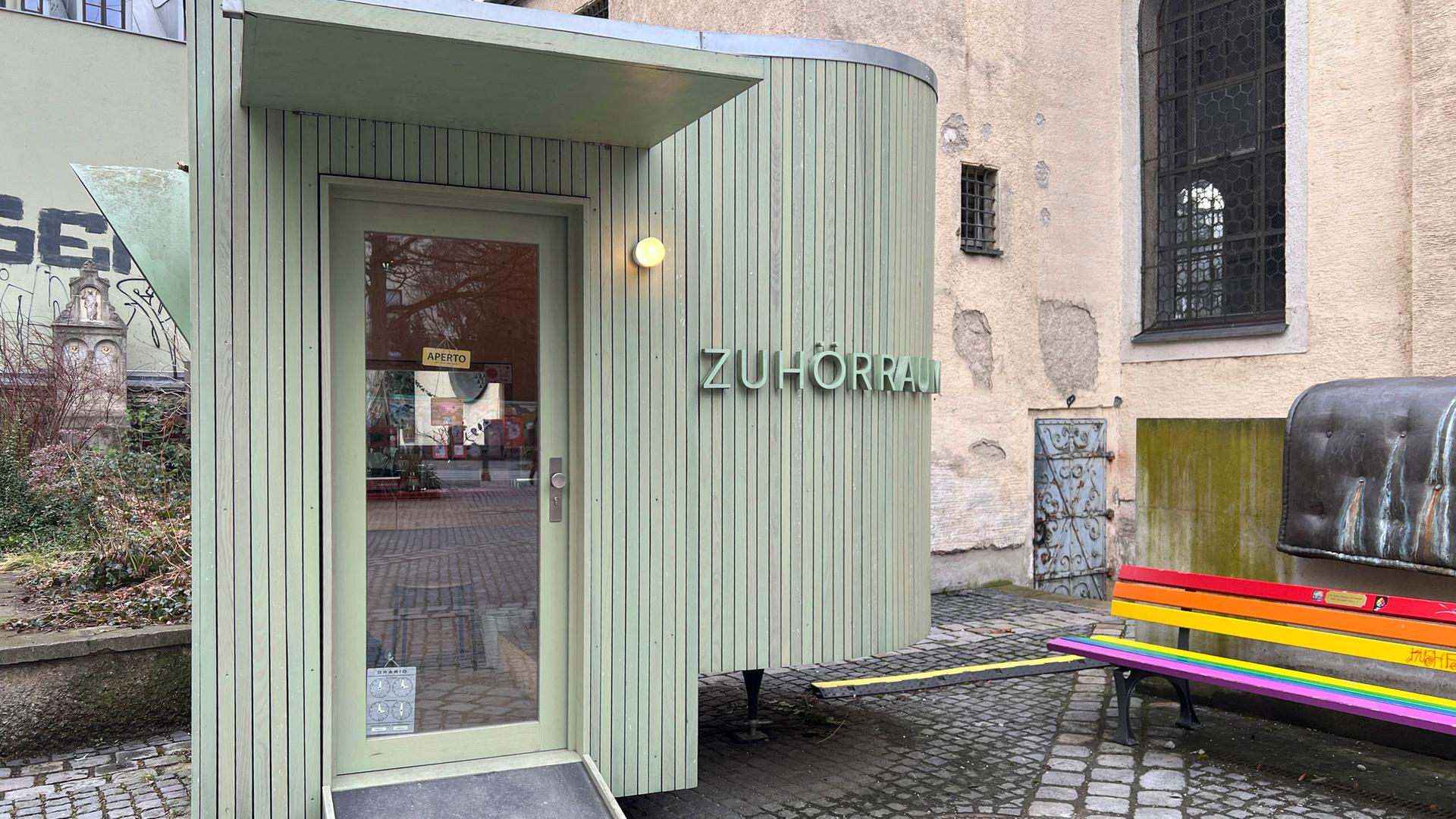 An einem oval geformten lindgrünen Holzbau neben der kleinen Kirche am Alten Südfriedhof in München steht "Zuhörraum". An einem oval geformten lindgrünen Holzbau neben der kleinen Kirche am Alten Südfriedhof in München steht "Zuhörraum".