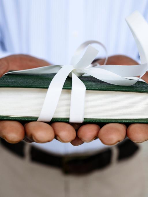 Nahaufnahme zweier Hände, die der Kamera ein mit weißem Geschenkband verziertes Buch entgegenhalten.