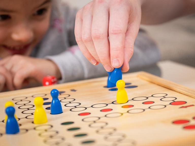 Nahaufnahme: Eine Kinderhand hält eine Spielfigur und schlägt damit eine andere aus dem Spiel "Mensch ärgere dich nicht" heraus, im Hintergrund sieht ein Mädchen gespannt zu und lacht.