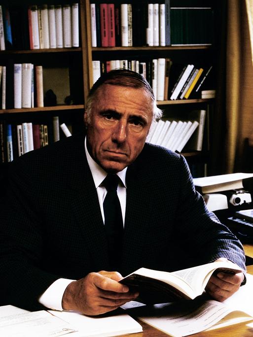 Der Suhrkamp-Verleger Siegfried Unseld sitzt an einem Schreibtisch, ein Buch in der Hand, und schaut in die Kamera. Hinter ihm steht ein gefülltes Bücherregal.