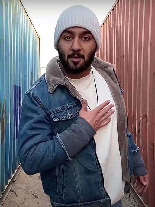 Der iranische Rapper Toomaj Salehi steht mit Wollmütze und Jeansjacke zwischen zwei Containern und fasst sich ans Herz.