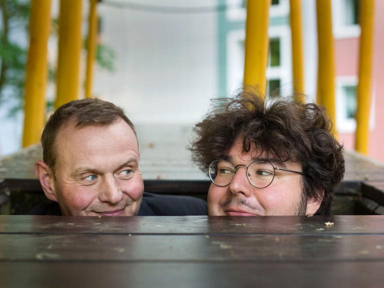 Devid Striesow und Axel Ranisch schauen aus einer hölzernen Bodenluke hervor und schauen sich verschmitzt an