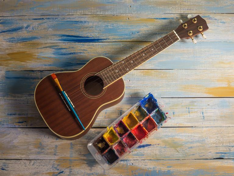 Eine Ukulele liegt neben einer Farbpalette, auf ihr sieht man einen Pinsel. 
